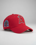 A hand holds a red Los Angeles Angels baseball cap with a white and red embroidered 'A' logo and a circular patch on the side, with other caps visible in a retail store background.